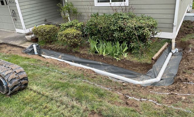 A trench lined with black fabric and white pipe for drainage near a house and garden.