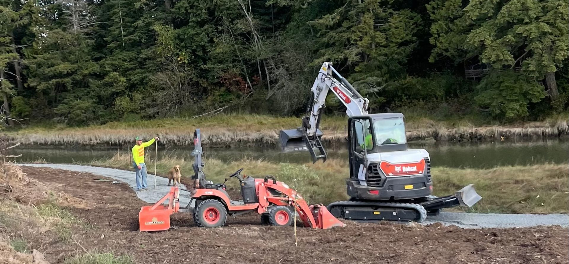 Construction site. A person in yellow stands near a tractor and excavator working beside a waterway with trees.