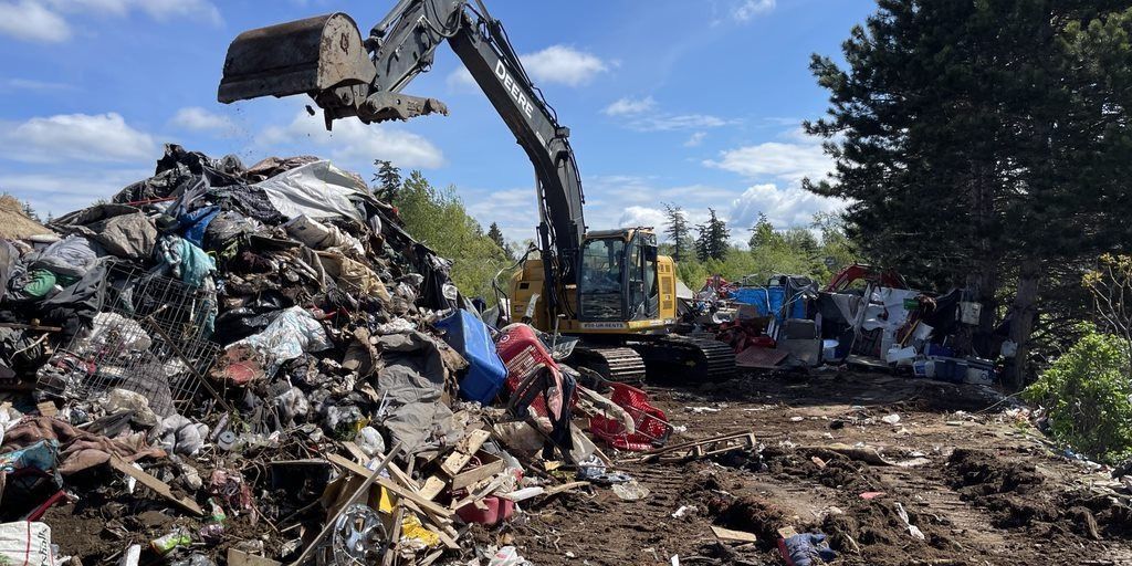 An excavator moving a pile of trash in a wooded area. The sky is blue and there are trees in the background.