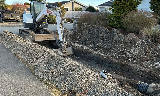 Mini excavator digging trench in residential area, gravel and dirt piles present.