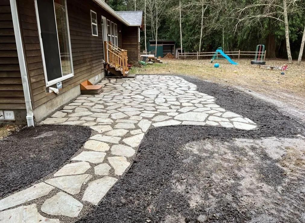 A stone patio and walkway alongside a wood-sided house, surrounded by dark mulch and a yard with play equipment.