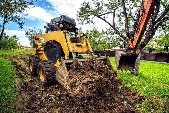 Yellow skid steer moving dirt on a green lawn with an orange excavator in the background.