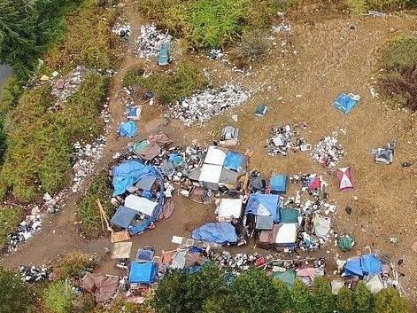 Overhead view of a makeshift encampment with tents, tarps, and trash scattered across a dirt area surrounded by vegetation.