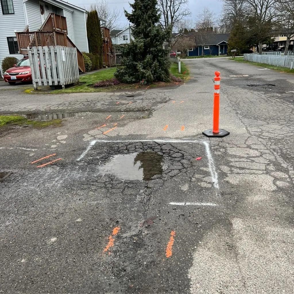 Pothole in asphalt road, marked with white and orange paint, puddle reflecting overcast sky; orange traffic cone.