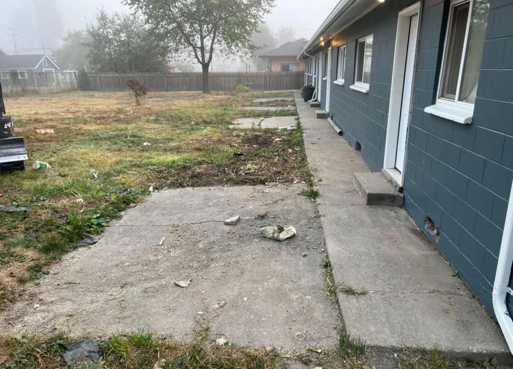 Exterior of a building with a concrete walkway and patches of concrete slabs in a grassy yard under a foggy sky.