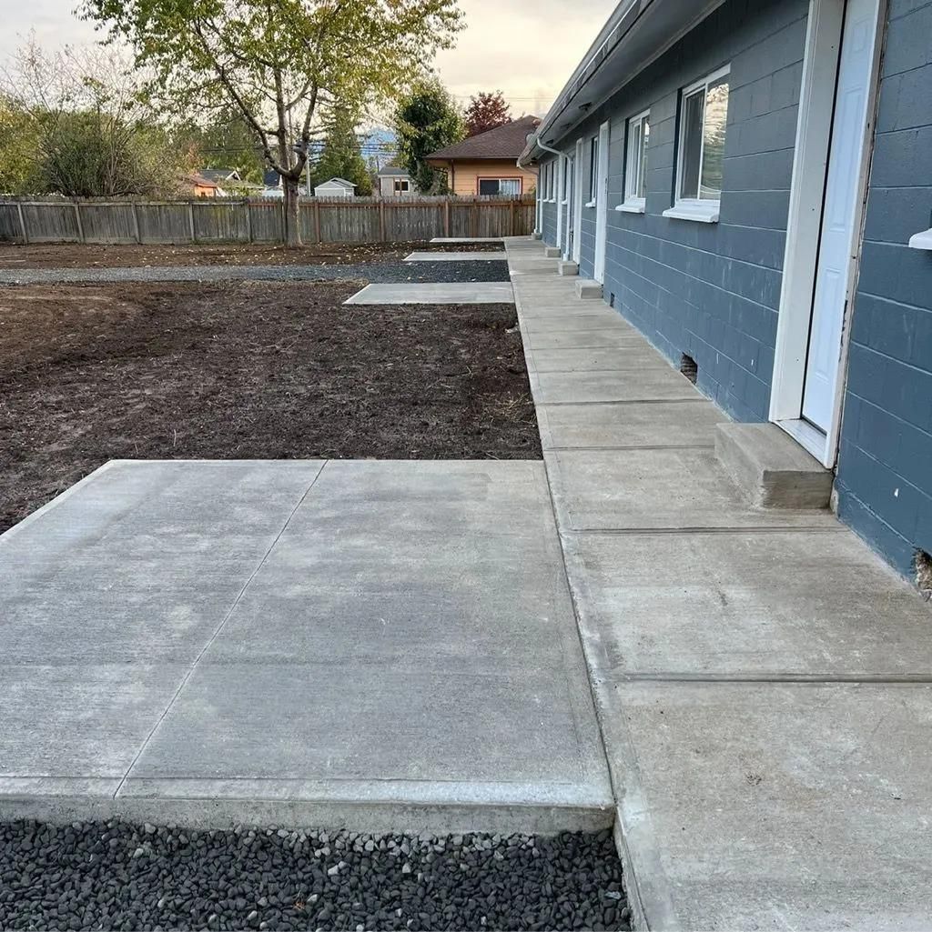 Concrete sidewalk and patios next to a blue building with white trim. Yard in background.
