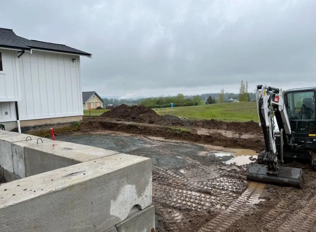 Construction site: Excavation, concrete foundation, small excavator near a white house. Cloudy day.