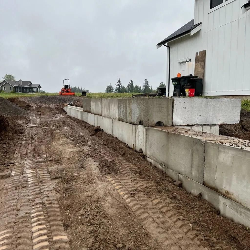 Construction site with concrete blocks forming a retaining wall, muddy ground, a building, and a roller compactor.