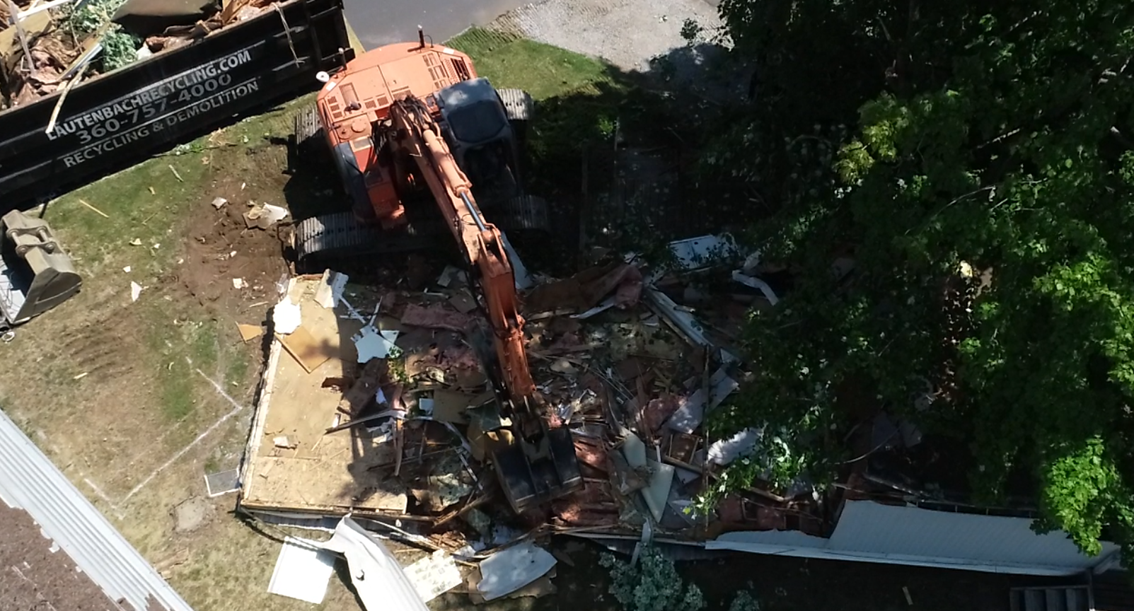 Orange excavator demolishing a building on a residential lot, debris visible.