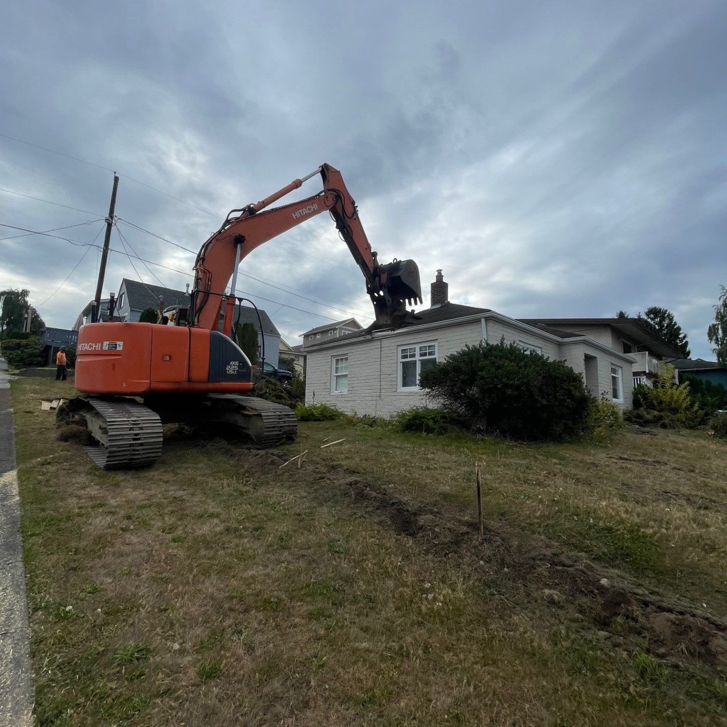 Excavator demolishing a one-story white house on a cloudy day.