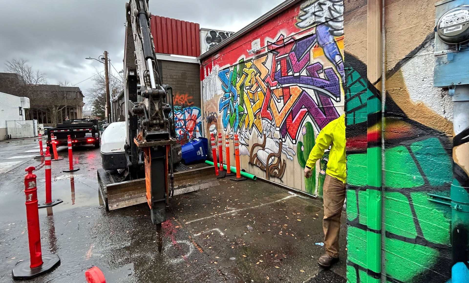 An excavator drilling pavement next to a colorful mural; a person in a yellow jacket stands nearby.