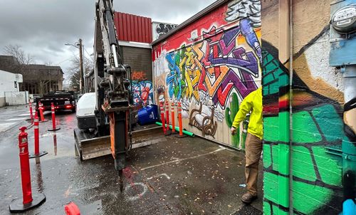 An excavator drilling pavement next to a colorful mural; a person in a yellow jacket stands nearby.