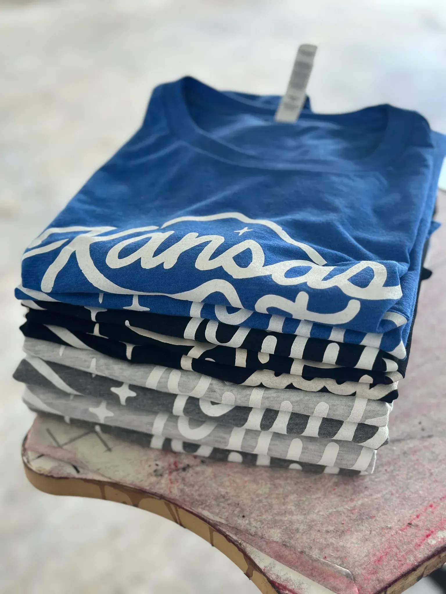 Stack of folded t-shirts; blue, black, and grey. The top shirt is blue with