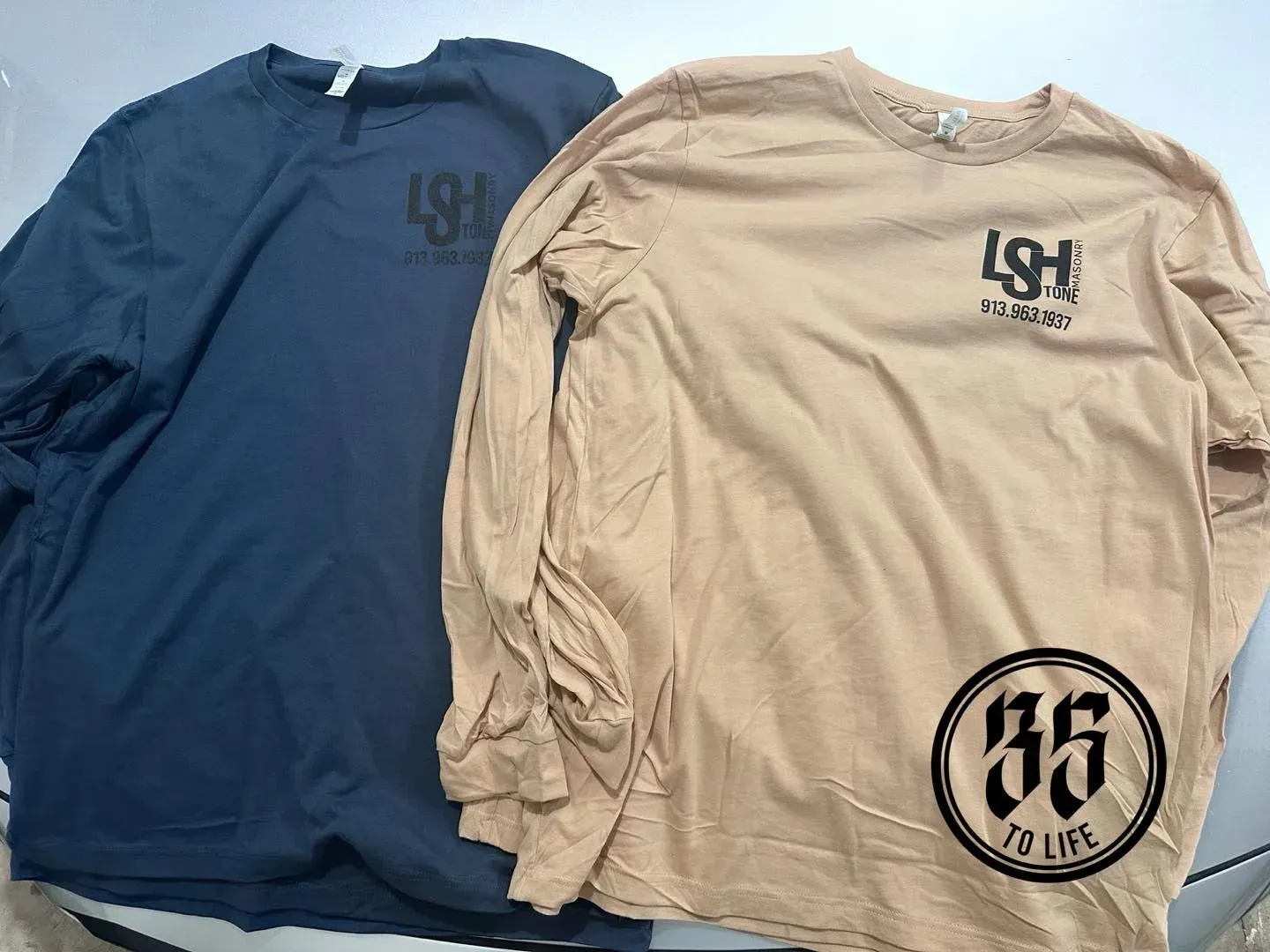 Two long-sleeved shirts: navy and tan. Both have a logo on the chest and a circled 