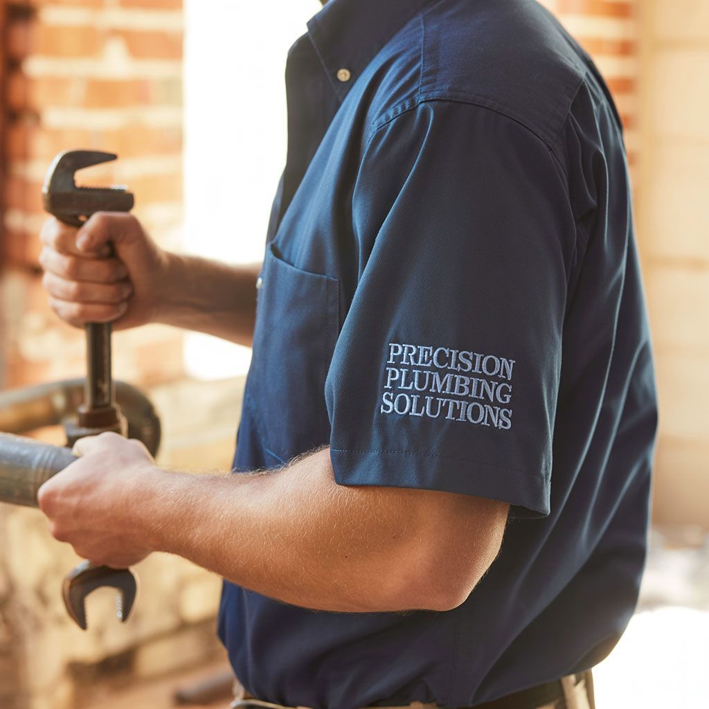 Plumber holding a wrench, wearing a navy blue shirt with company logo, working near brick wall.