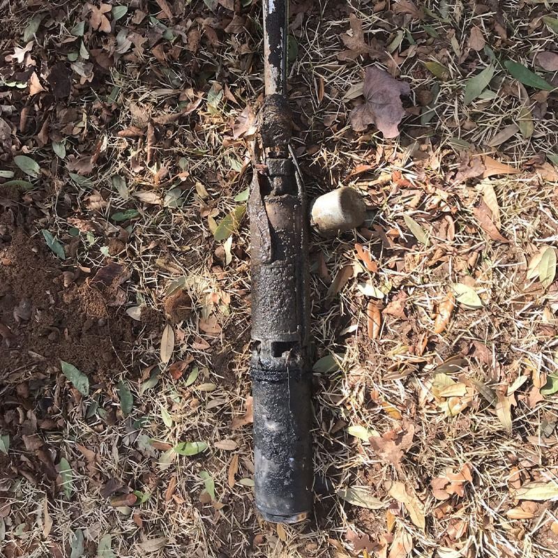 Rusted well pump laying on the ground among fallen leaves and brown soil.