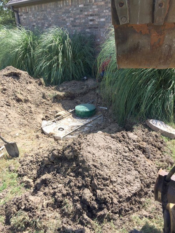 Excavated septic tank area, green lid visible. Dirt pile, grass, and tall plants in background.