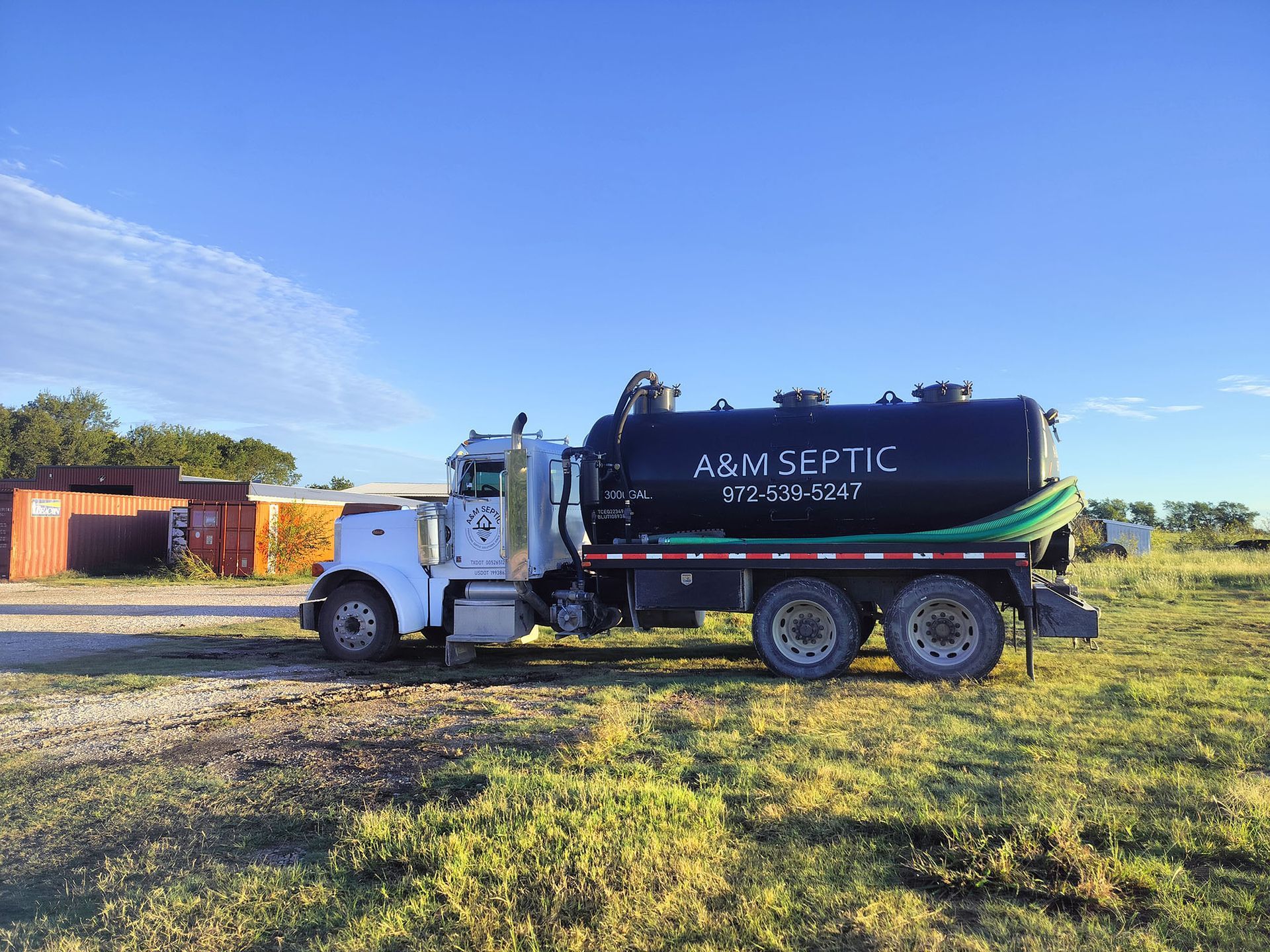 White septic truck parked on a grassy field under a blue sky. The truck's tank is black with company lettering.
