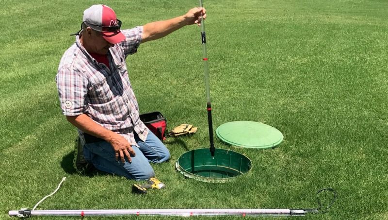 Man kneeling, measuring depth of a green septic tank access point in a grassy yard.
