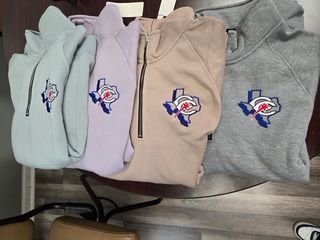 Four folded hoodies in mint, lavender, beige, and gray with sports-style logos on a shelf