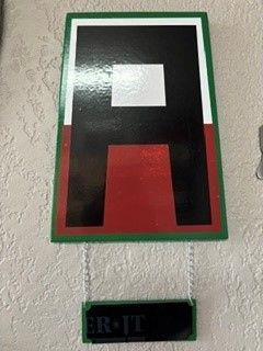 Abstract black, red, and white wall sign with a green border, hanging above a small label.