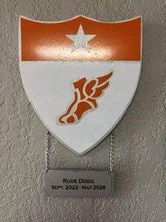 Shield-shaped plaque with orange top, white star and logo, labeled “Rude Dogg” and “Sept. 2022 - May 2026.”