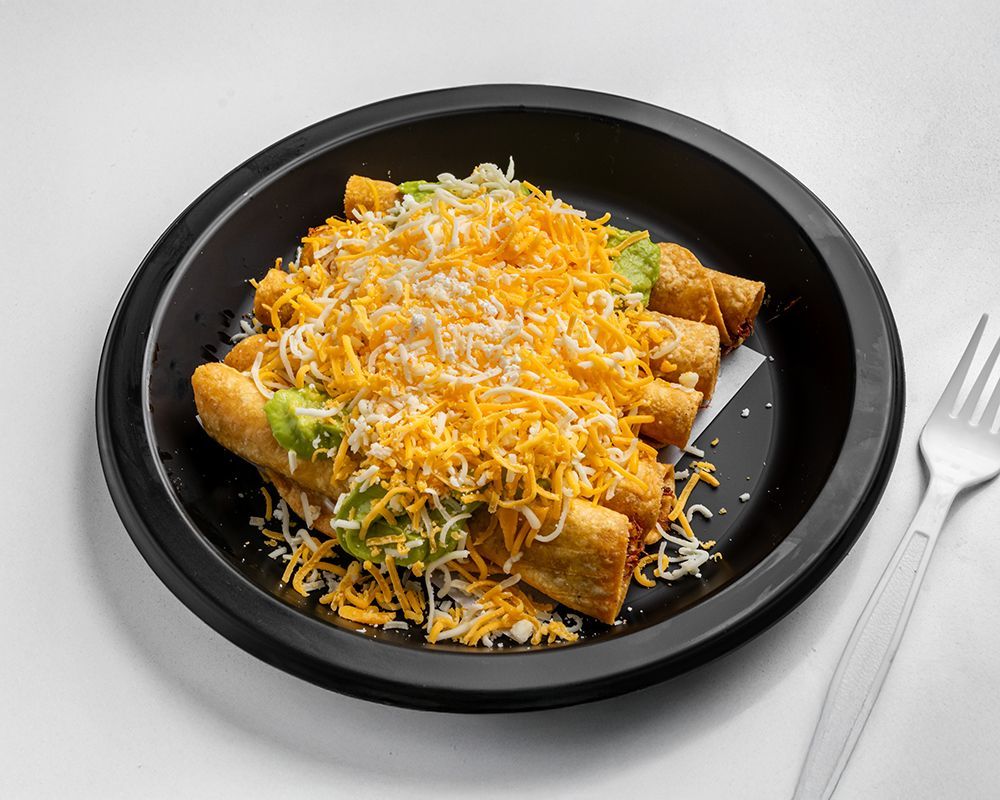 Rolled tacos topped with cheese and guacamole on a black plate, next to a fork.