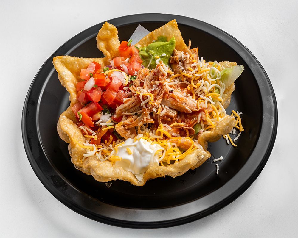 Taco salad in a fried tortilla bowl with chicken, vegetables, and toppings, on a black plate.