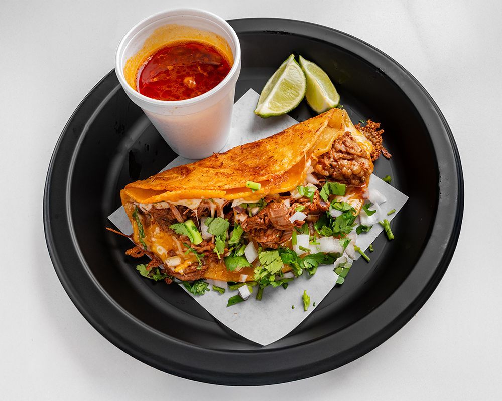 Quesabirria taco with dipping sauce and lime wedges on a black plate.