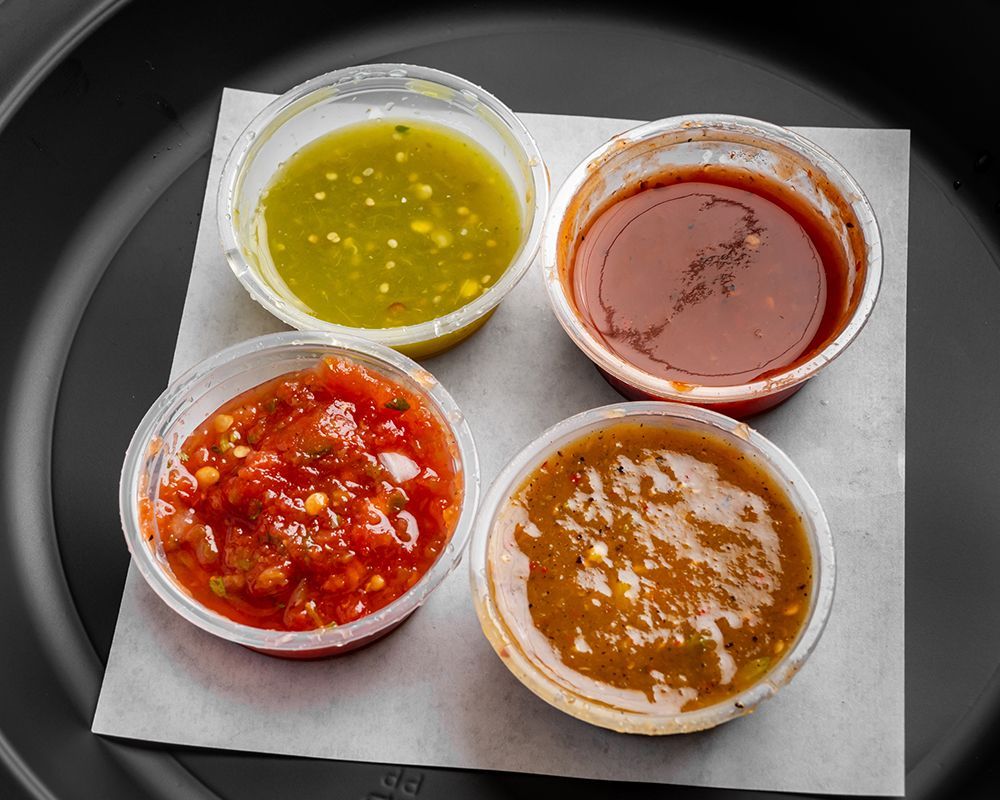 Four small plastic cups of salsa, with various colors and textures, on a white square.