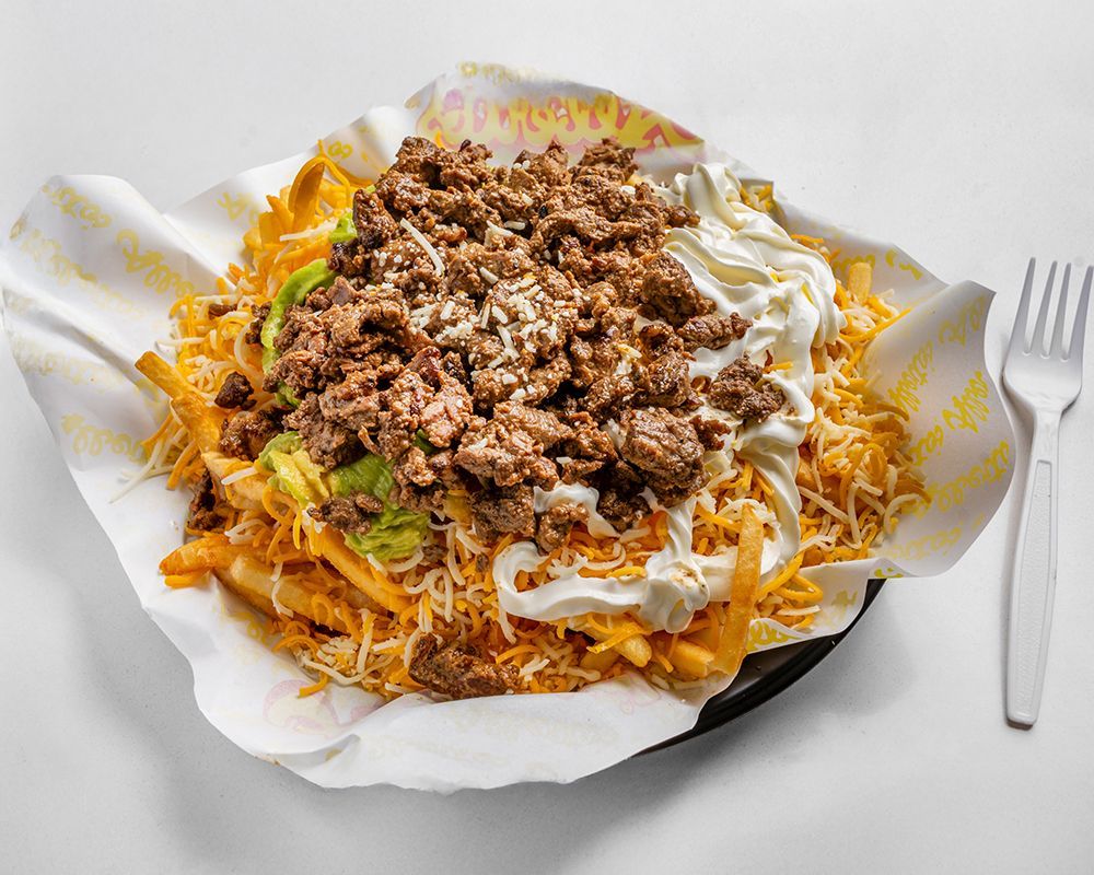 Loaded fries with ground meat, cheese, sour cream, and guacamole, served on paper.