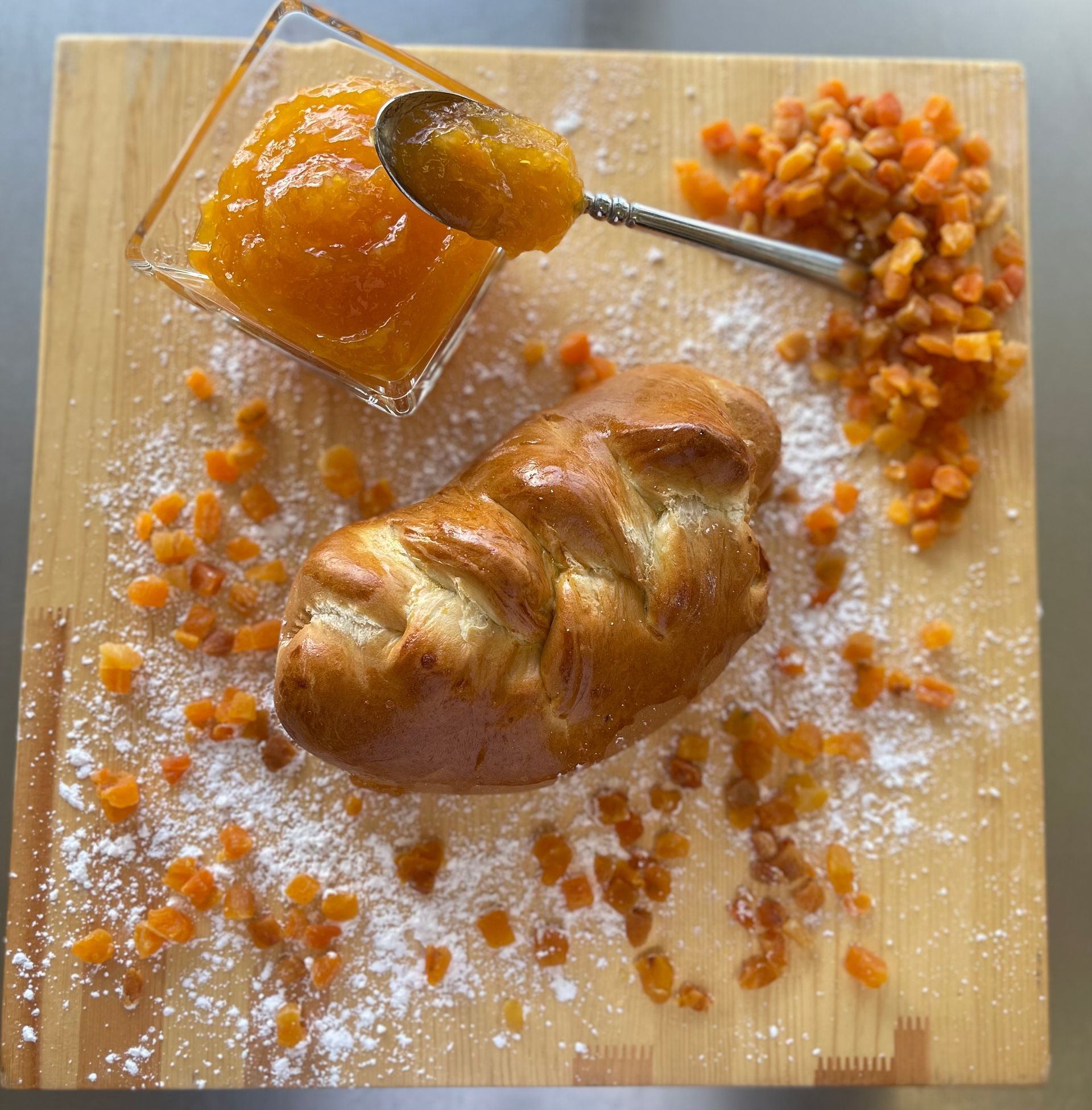 A loaf of bread is sitting on a wooden cutting board next to a bowl of jam and dried apricots.