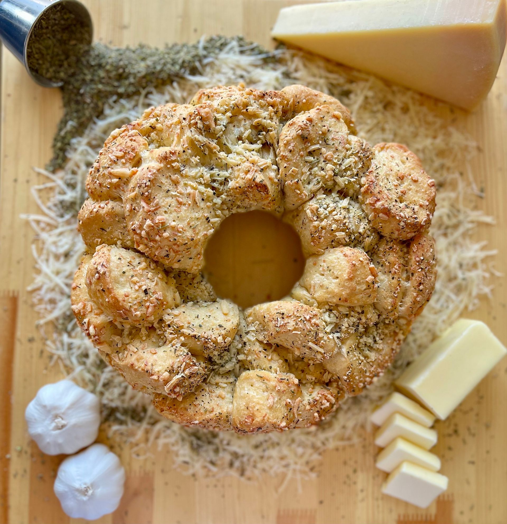 A bread wreath is sitting on top of a wooden cutting board next to garlic and cheese.
