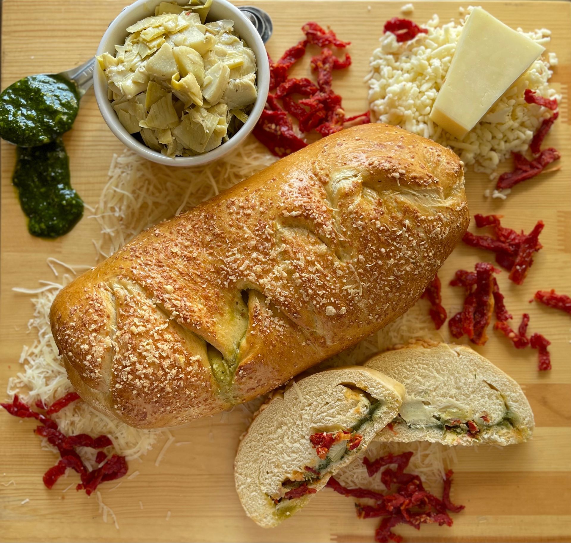 A loaf of bread is sitting on a wooden cutting board next to a bowl of artichokes and cheese.