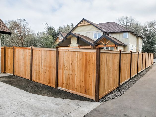 Wooden privacy fence around a two-story beige house with a brown roof; cloudy sky.