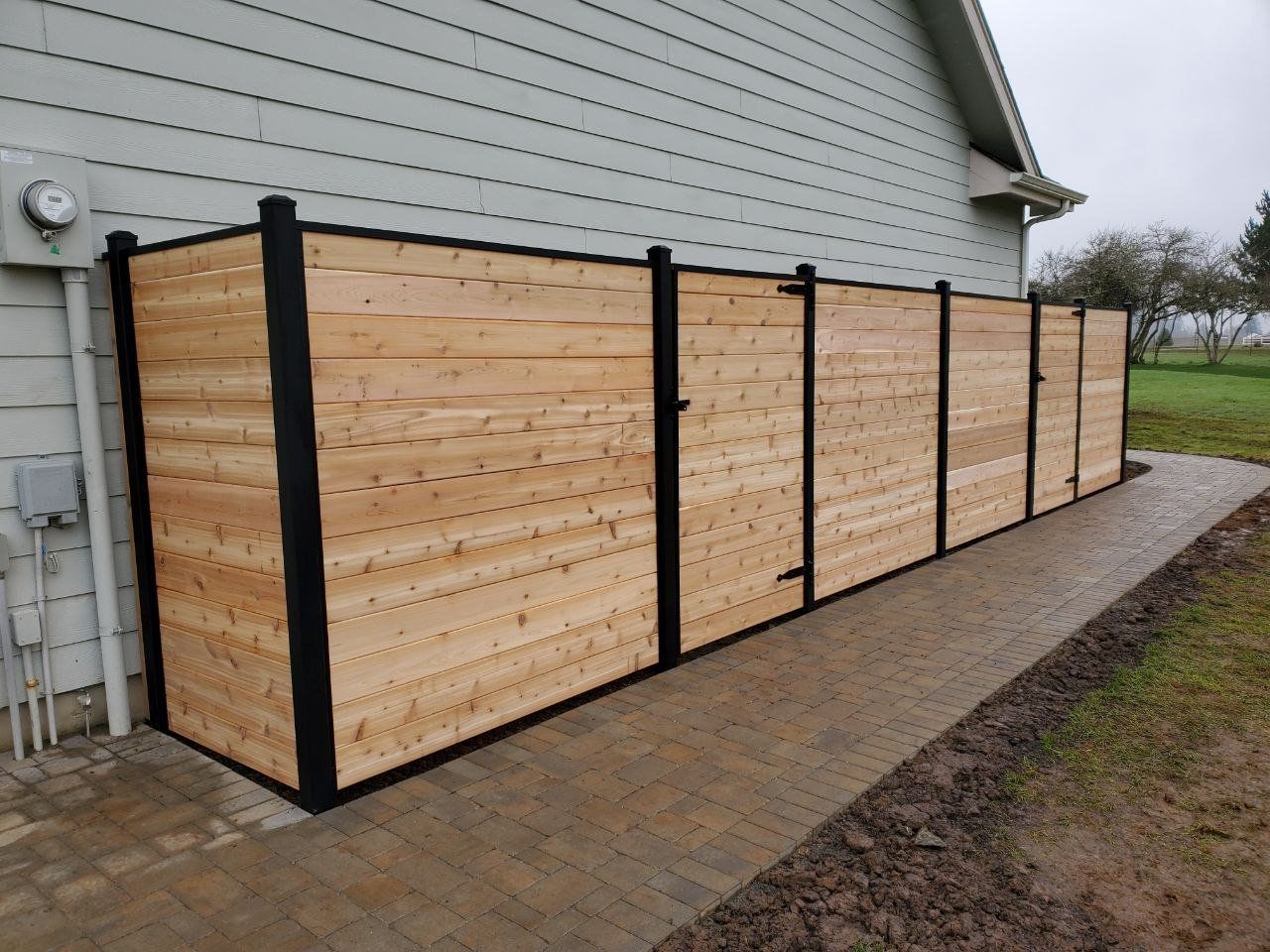 Wooden privacy fence along the side of a house, black posts, light brown planks, on a concrete path.