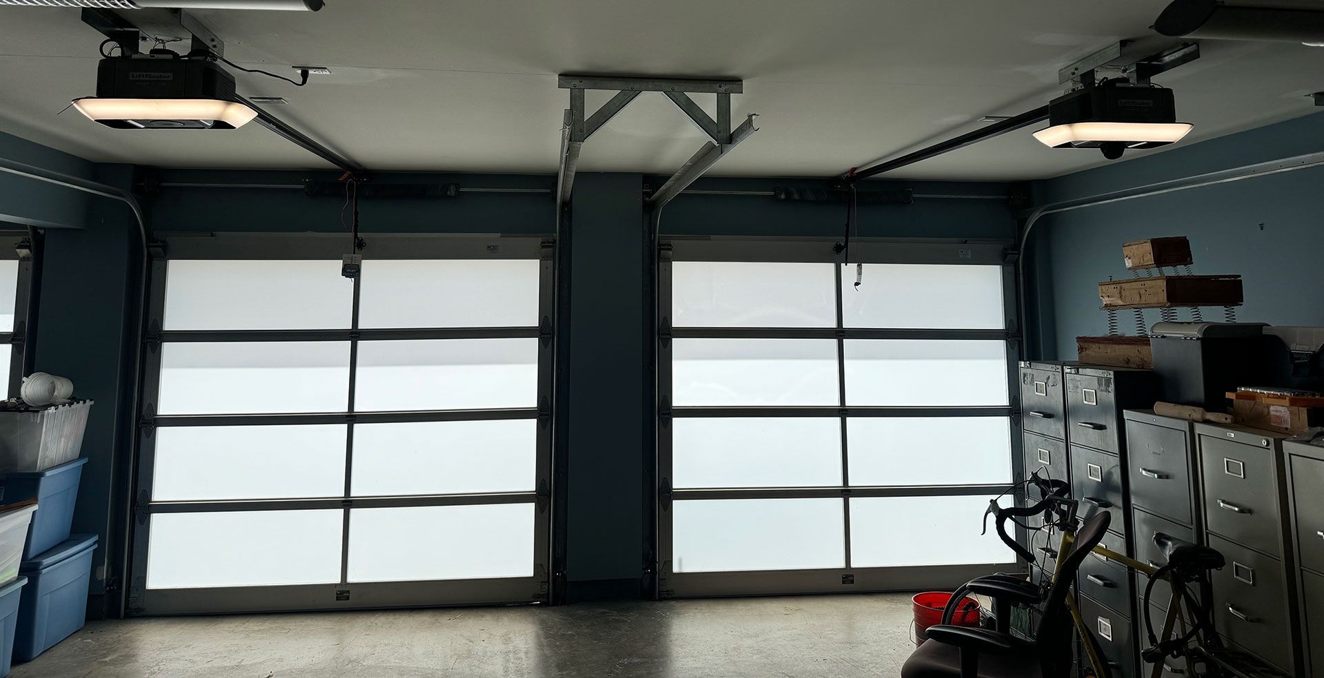 Two garage doors with glass panels and rolling shades, open inside a blue-walled garage.