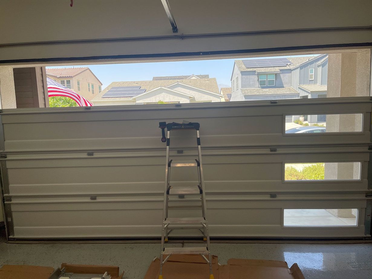 Garage door partially open with ladder inside, showing exterior homes and sky.