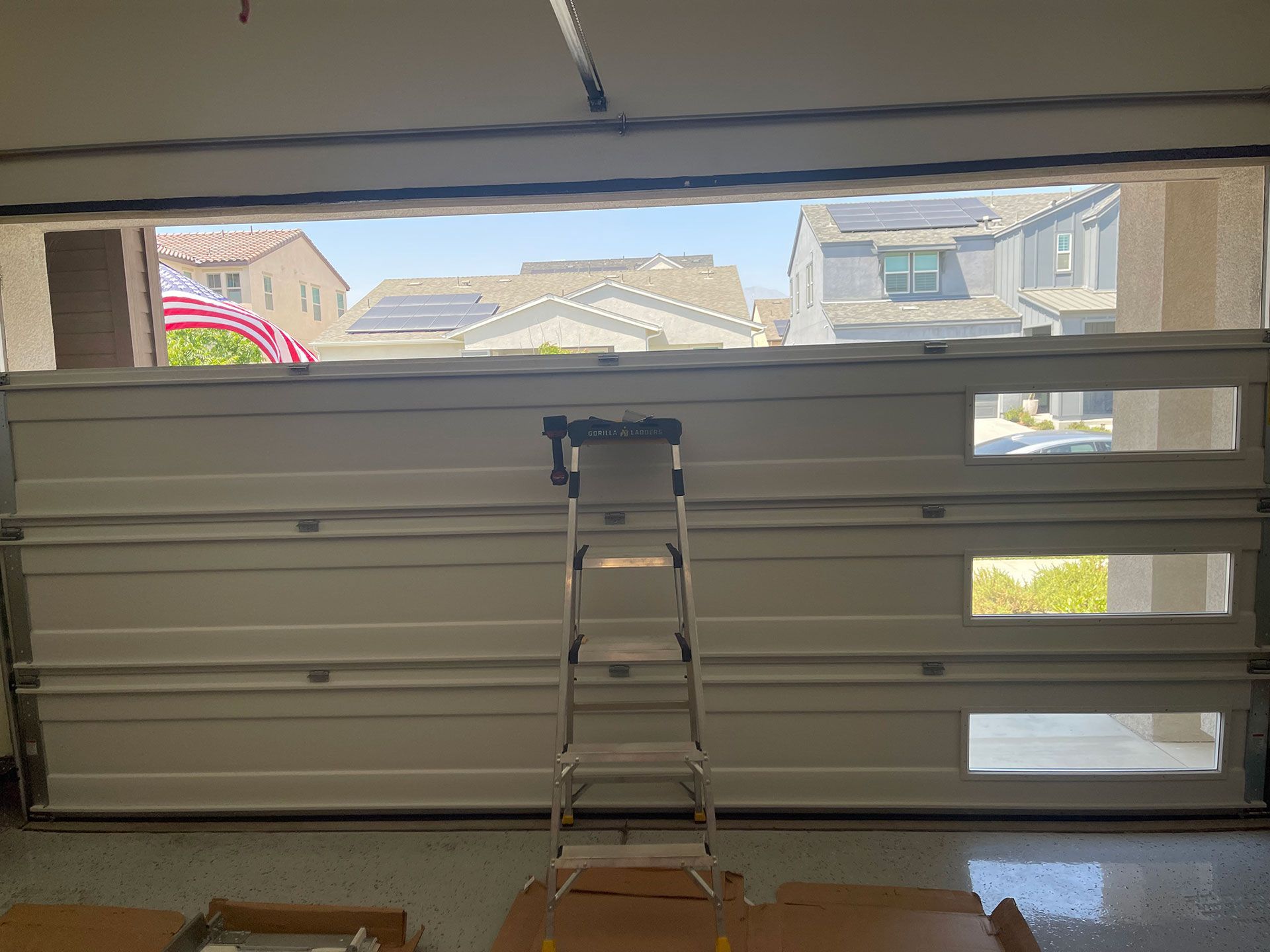 Garage door partially open with ladder inside, showing exterior homes and sky.