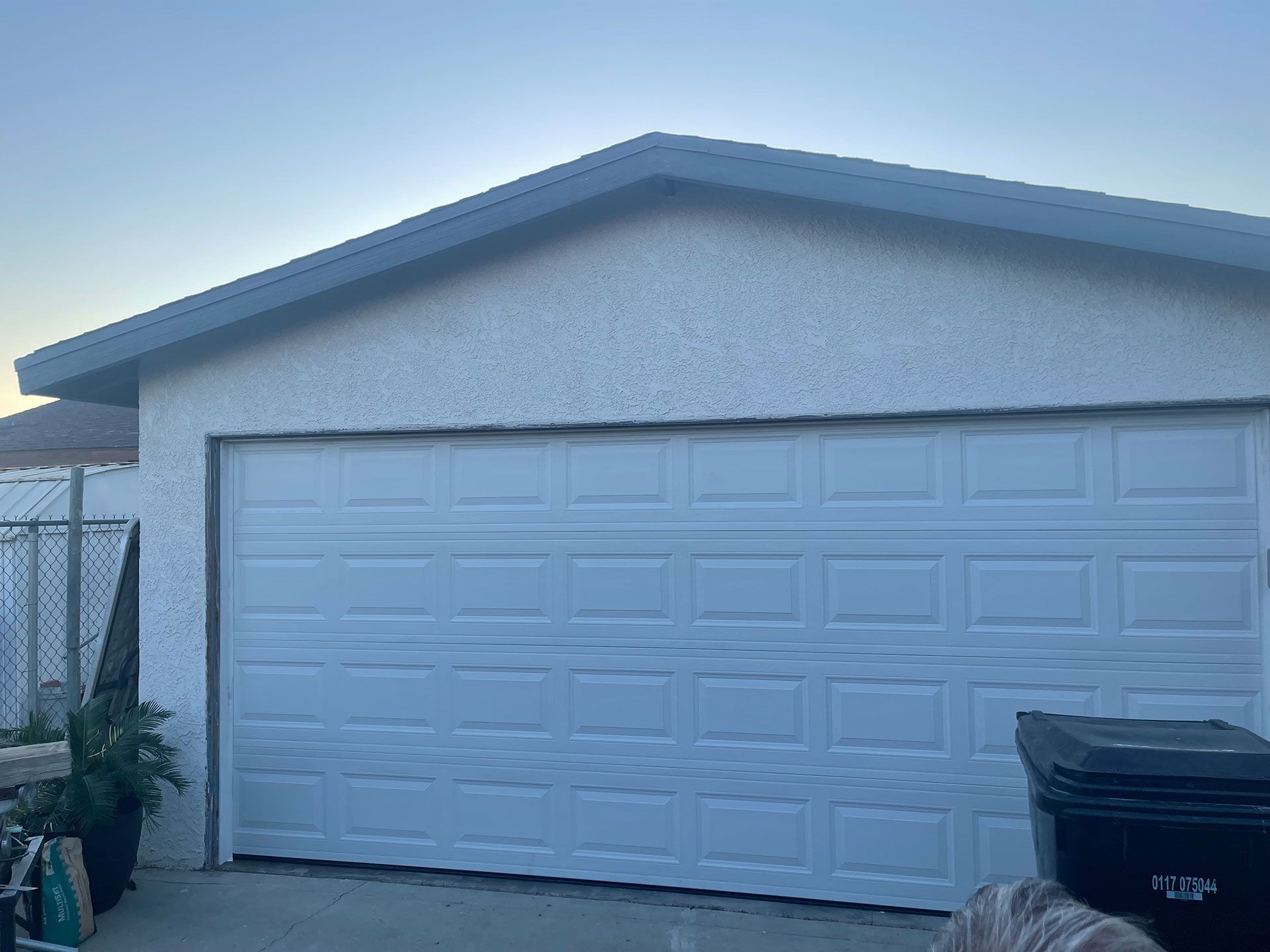 White garage door on a stucco building with a dark roof and a trash bin to the right.