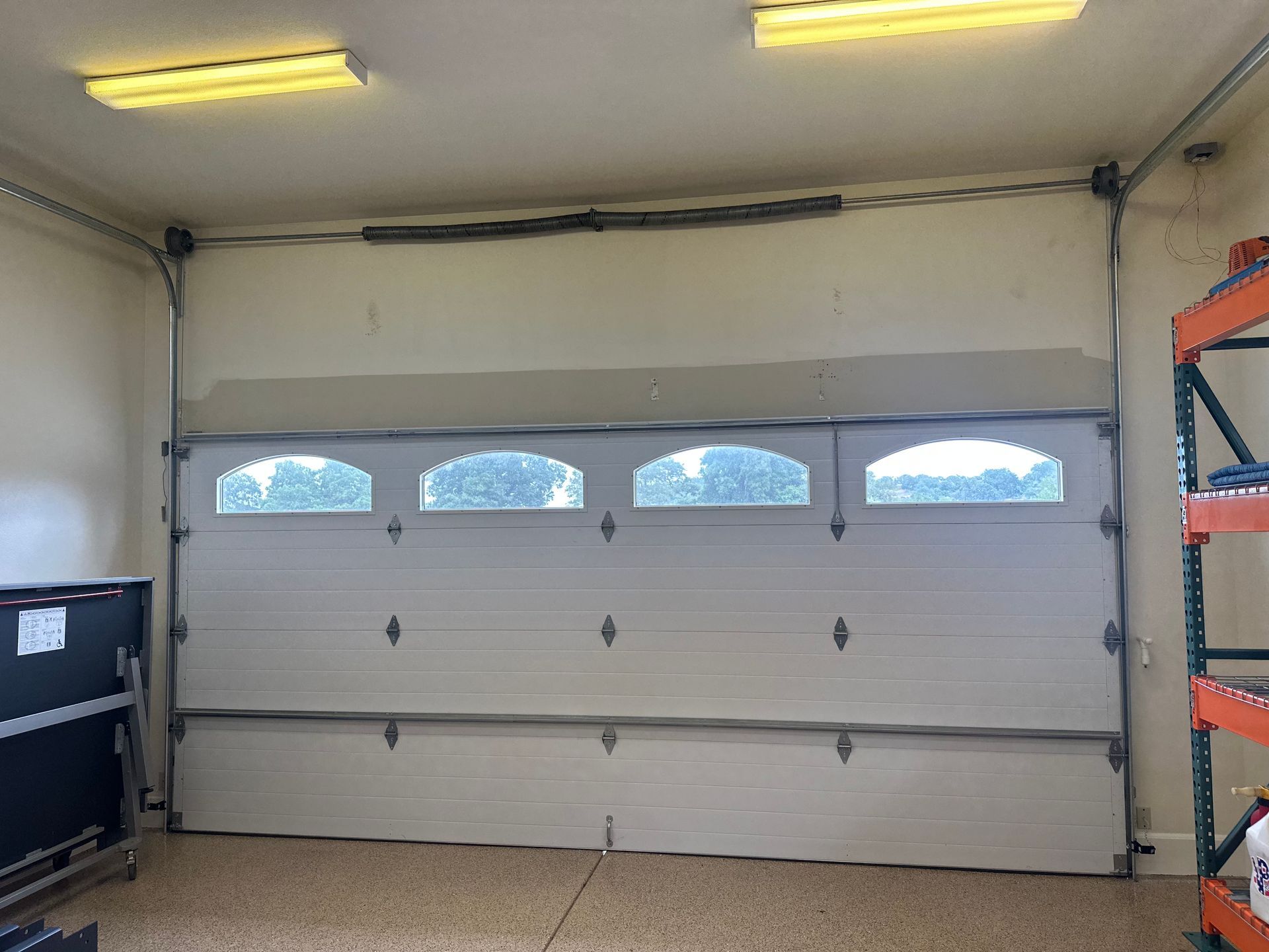 White garage door, partially open, with windows, in a room with a concrete floor and fluorescent lights.