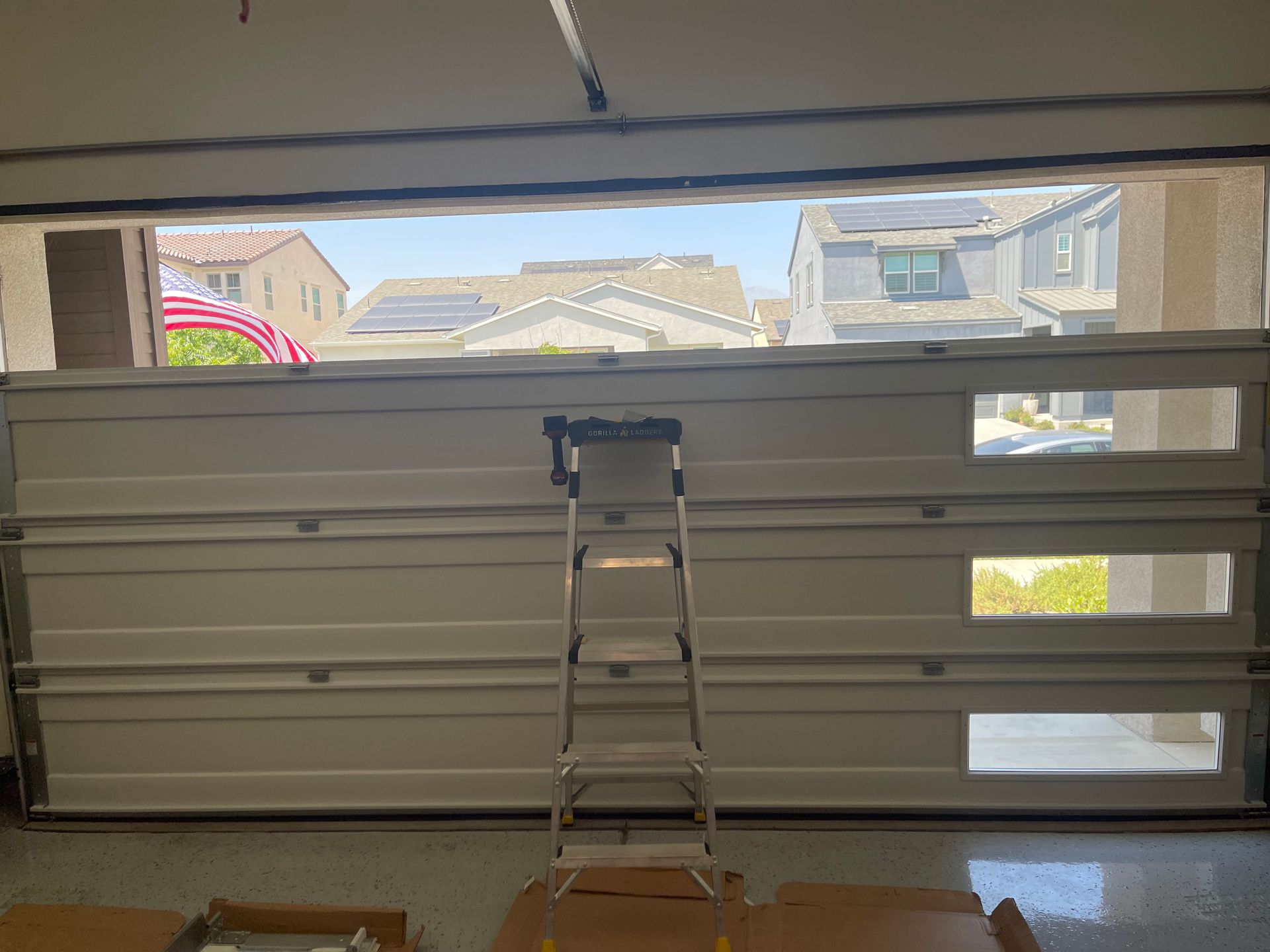 Interior view of a garage with an open door, a ladder, and a glimpse of houses outside.