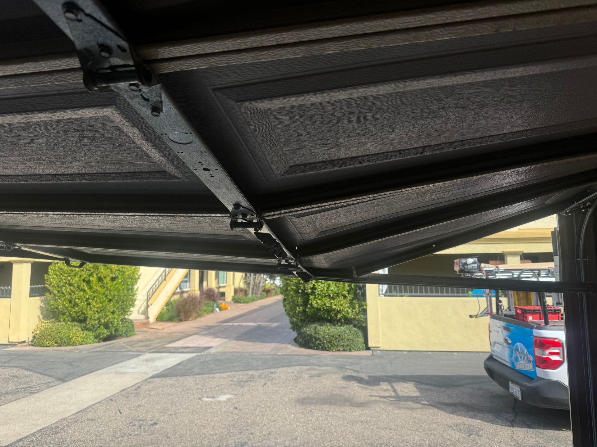Interior view of a garage door, partially open. The exterior view shows a street, greenery, and a vehicle.