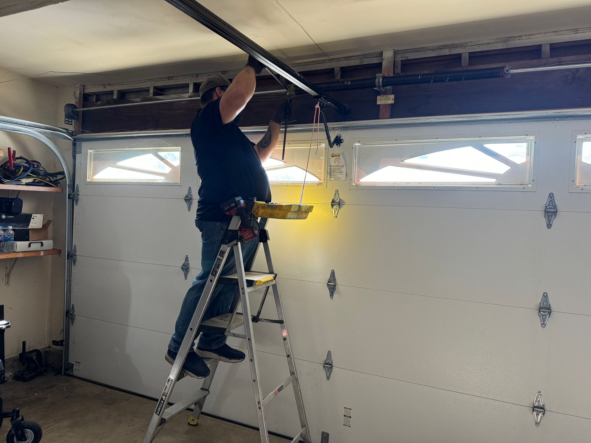 Person on stepladder installing garage door opener track above a closed garage door.