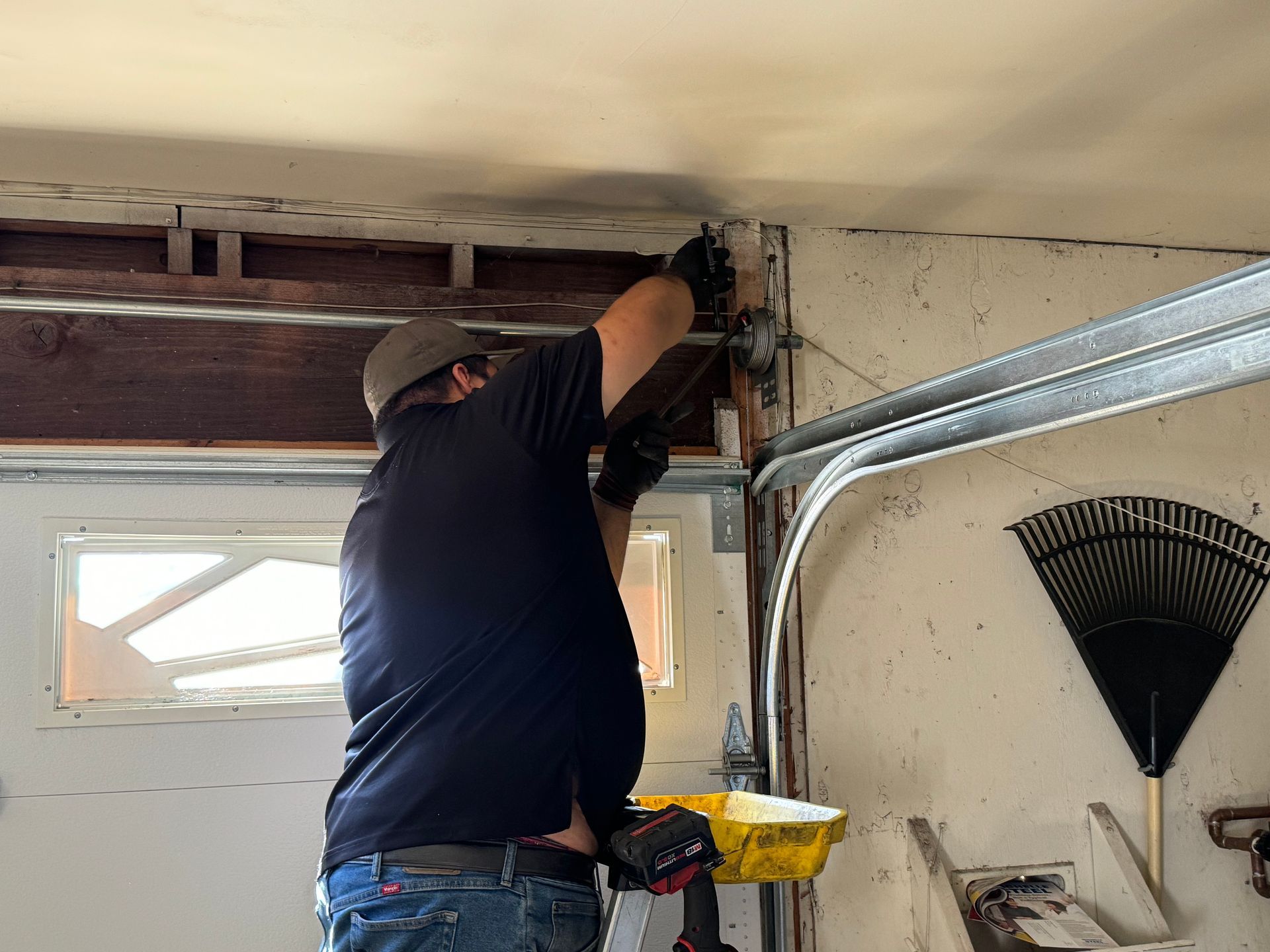 Person in a garage repairs overhead door mechanism, using tools. The garage door track and a rake are visible.