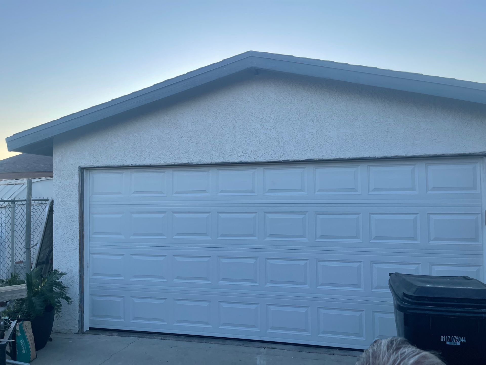 White garage with closed door, gray roof trim, blue sky. Black trash bin to the right.