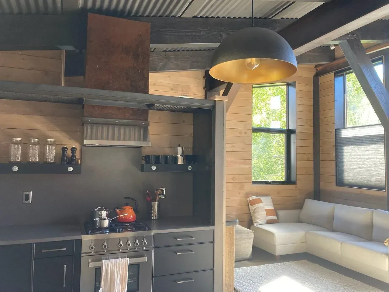 Rustic kitchen with a stove, black cabinets, and a gold-lined pendant light. A window lets in sunlight onto a cream-colored couch.