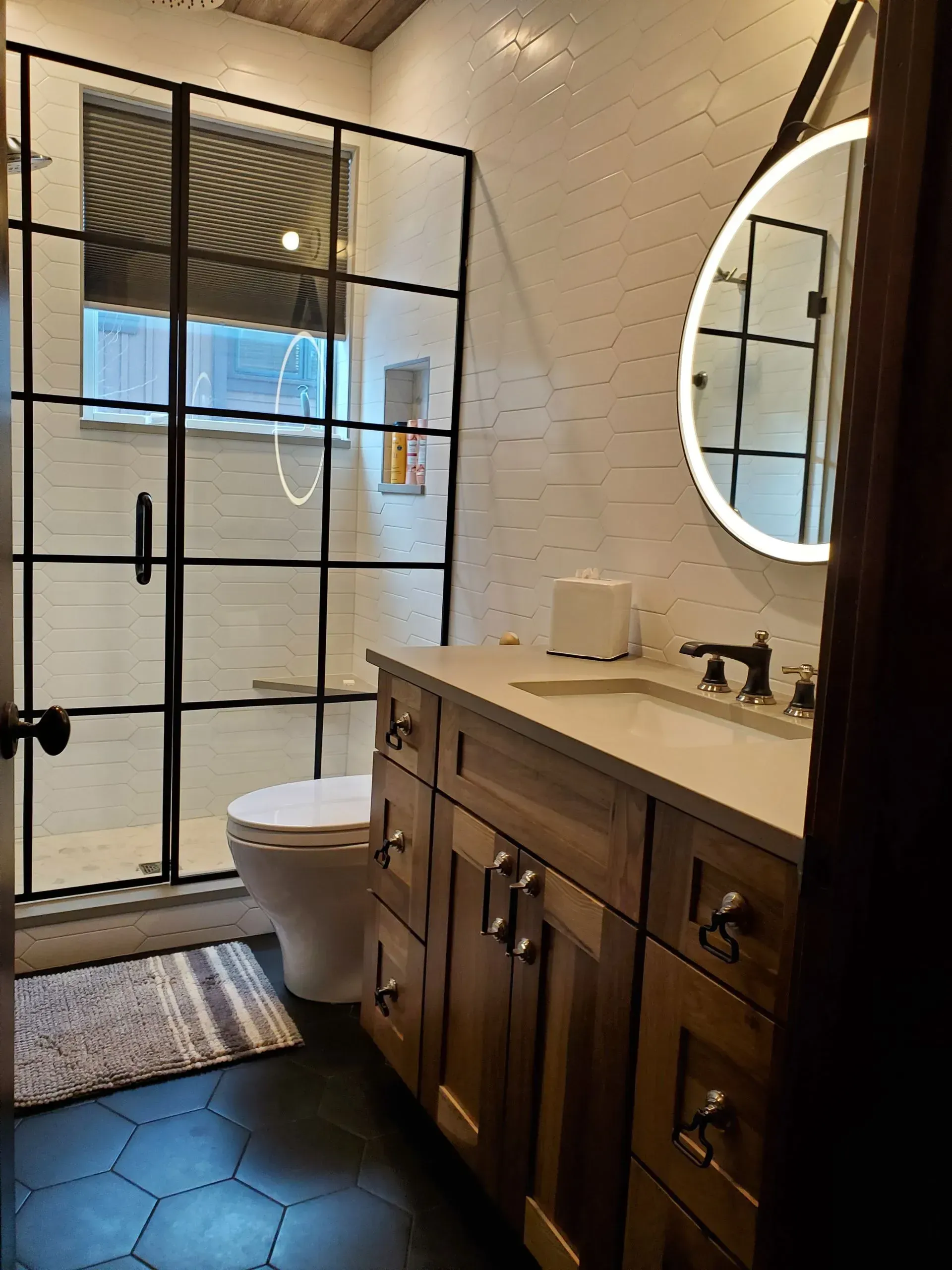 Bathroom with shower, vanity, toilet, and round mirror. Black-framed shower, wooden vanity, white tiled walls.