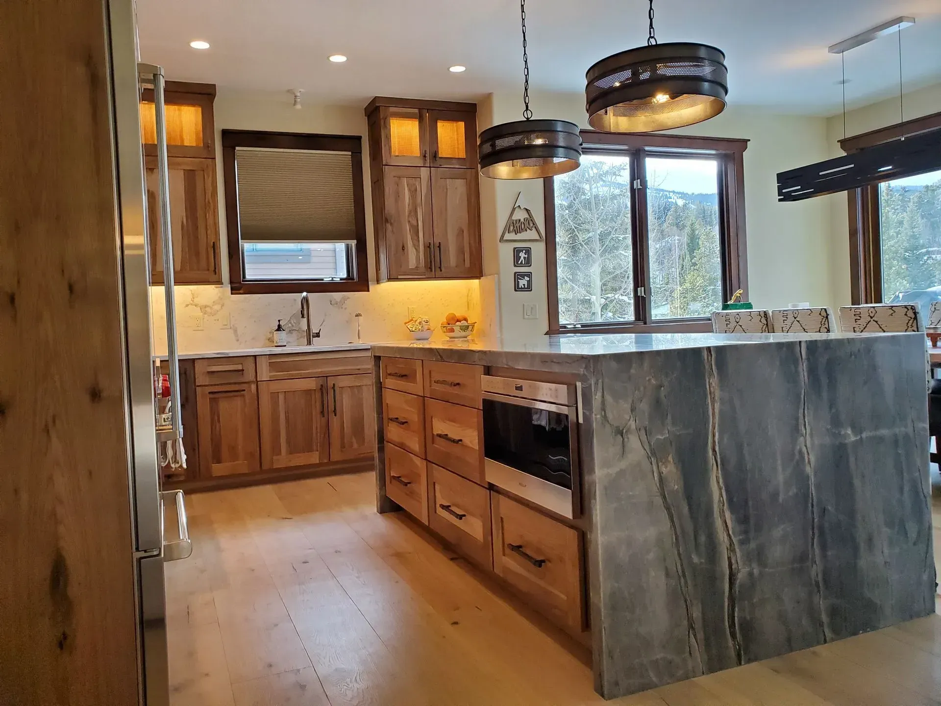 Kitchen with wooden cabinets, stone island, light wood floor, large window with a view.