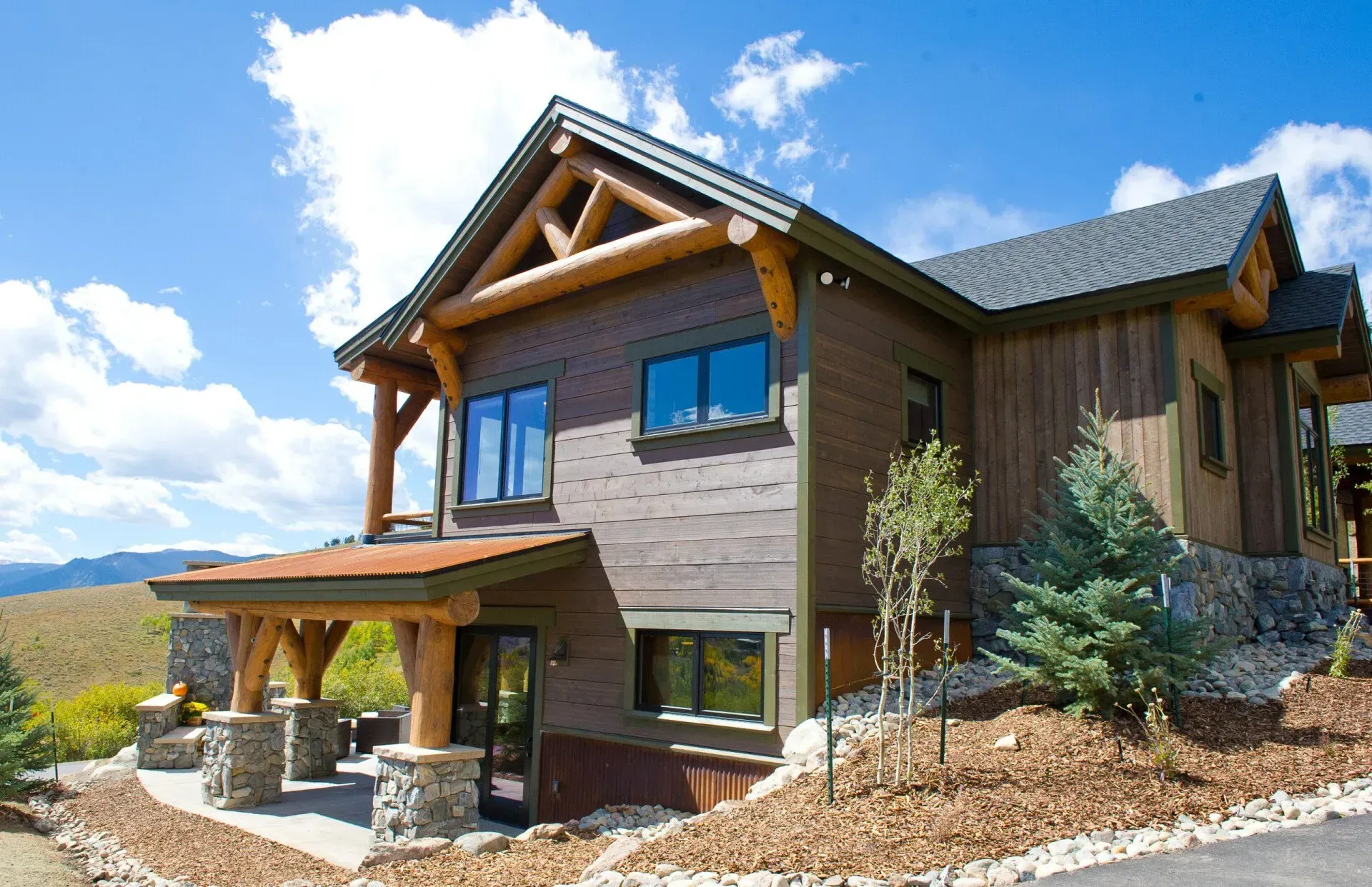 Two-story brown wood house with stone pillars, nestled in a hillside under a blue sky.